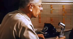 Photo of man sitting at desk with a phone and microphone in front of him.
